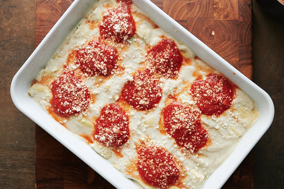 A baking dish filled with unbaked chicken cannelloni topped with white sauce, red tomato sauce, and grated cheese, placed on a wooden surface.