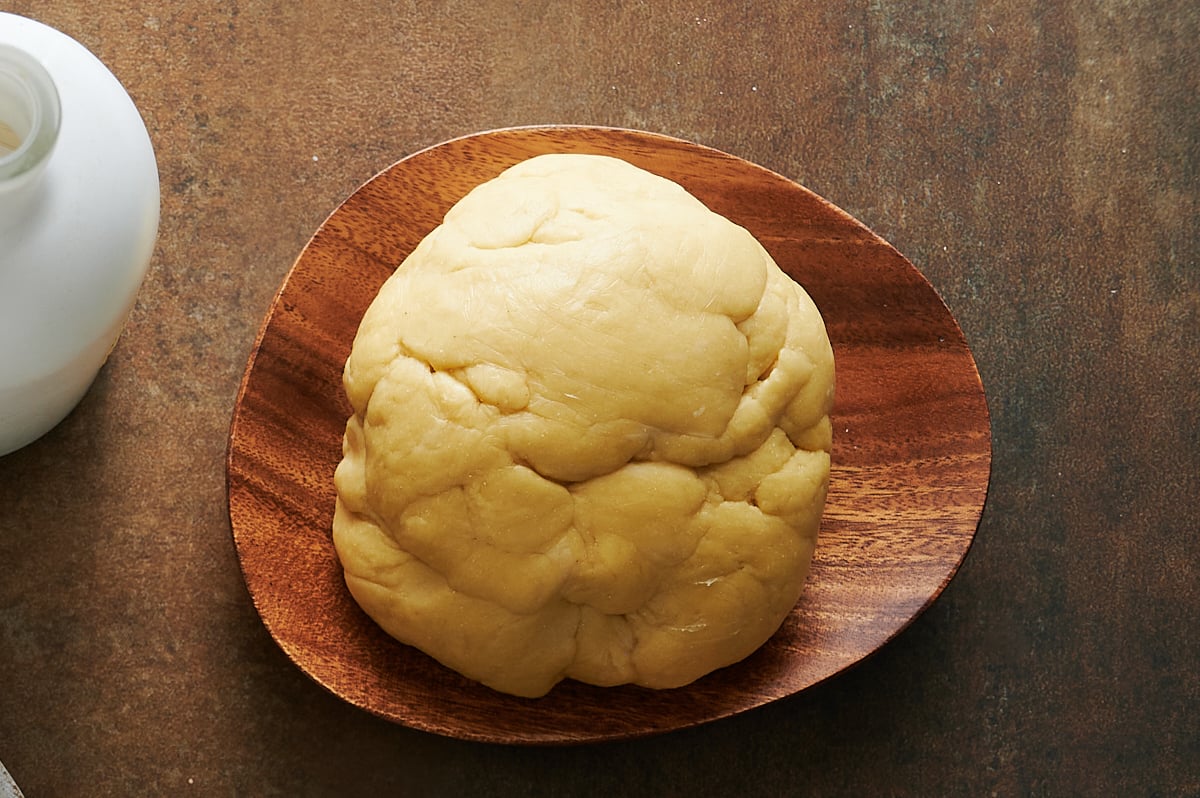 A ball of pasta dough sits on a wooden plate on a brown surface, with part of a bottle visible in the upper left corner.