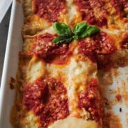 A baked dish of lasagna topped with tomato sauce, grated cheese, and a sprig of fresh basil in the center, shown in a white rectangular baking dish.