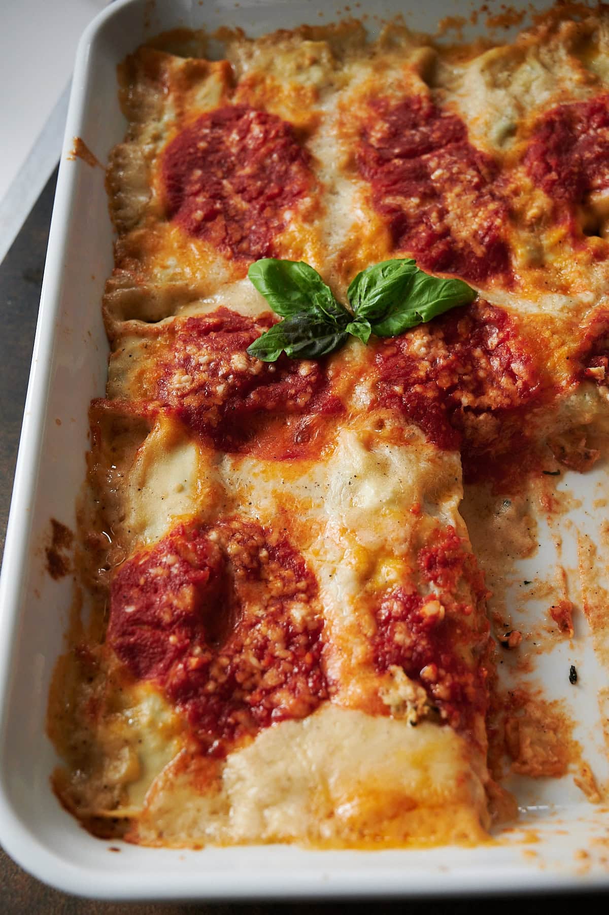 A baked dish of lasagna topped with tomato sauce, grated cheese, and a sprig of fresh basil in the center, shown in a white rectangular baking dish.