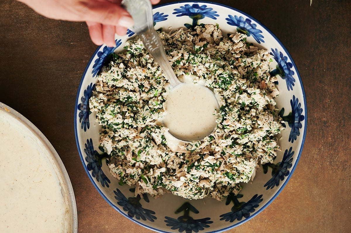 A hand pours a ladle of white sauce over a bowl of minced chicken and spinach mixture in a blue-and-white patterned dish.