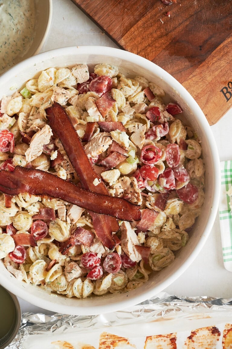A bowl of ranch pasta salad with cherry tomatoes, celery, chicken, and two strips of bacon on top, shown on a table next to a wooden cutting board and other food items.