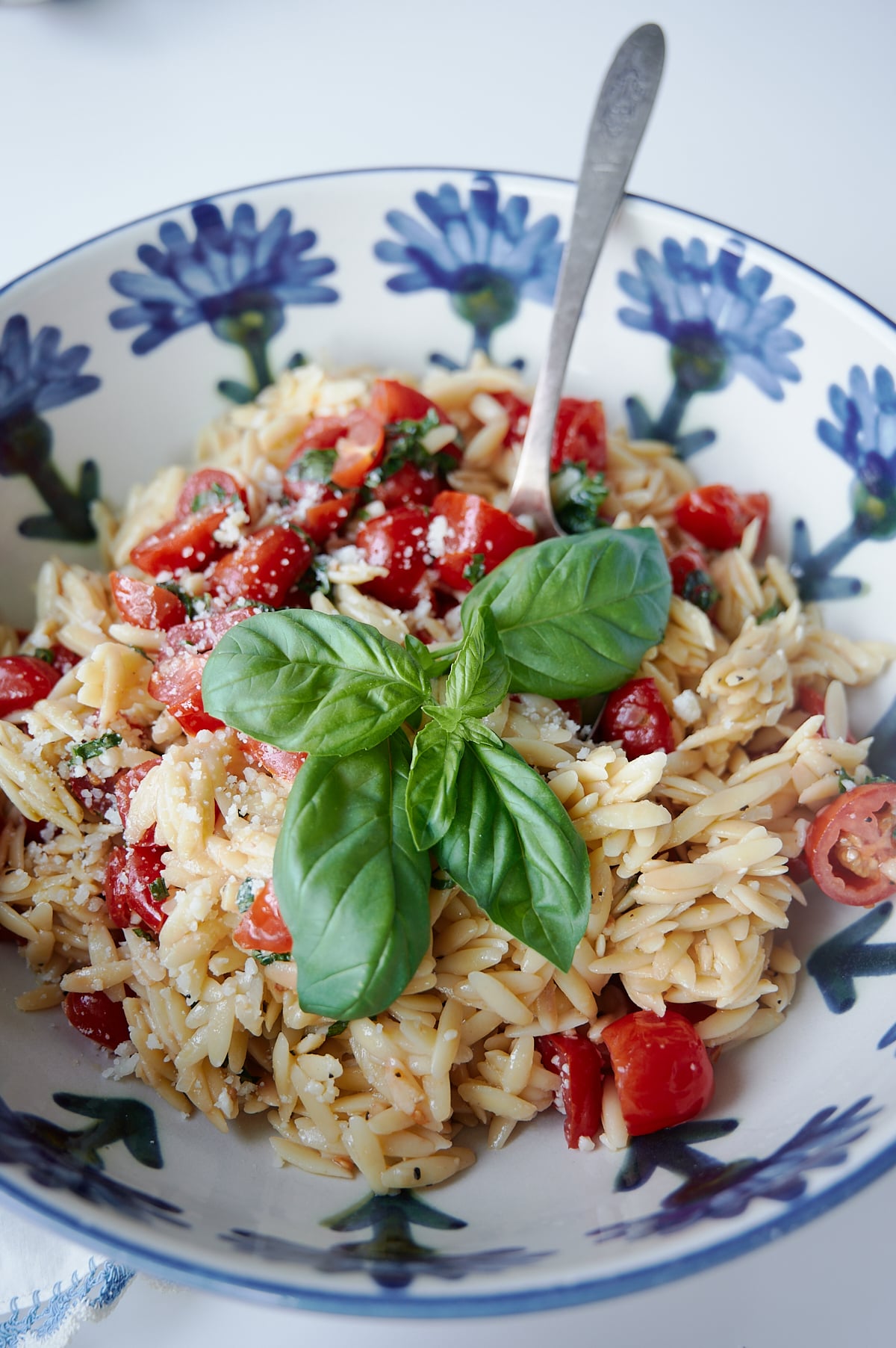 Orzo pasta with cherry tomatoes, grated cheese, garlic and fresh basil in a floral blue and white bowl with a spoon.