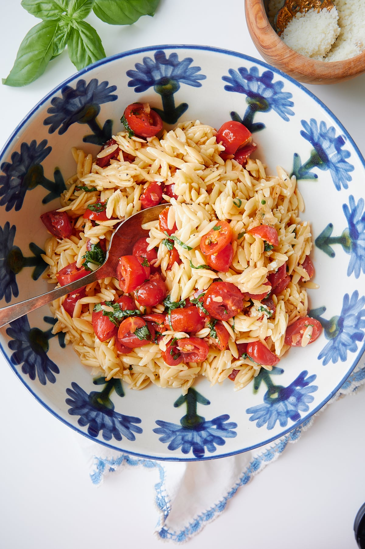 A floral bowl filled with orzo pasta tossed with halved cherry tomatoes and fresh basil, with a serving spoon resting inside.