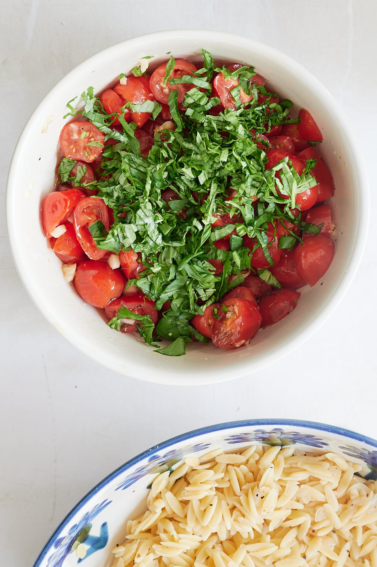 A bowl of halved cherry tomatoes and garlic topped with chopped basil, next to a bowl of cooked orzo pasta.