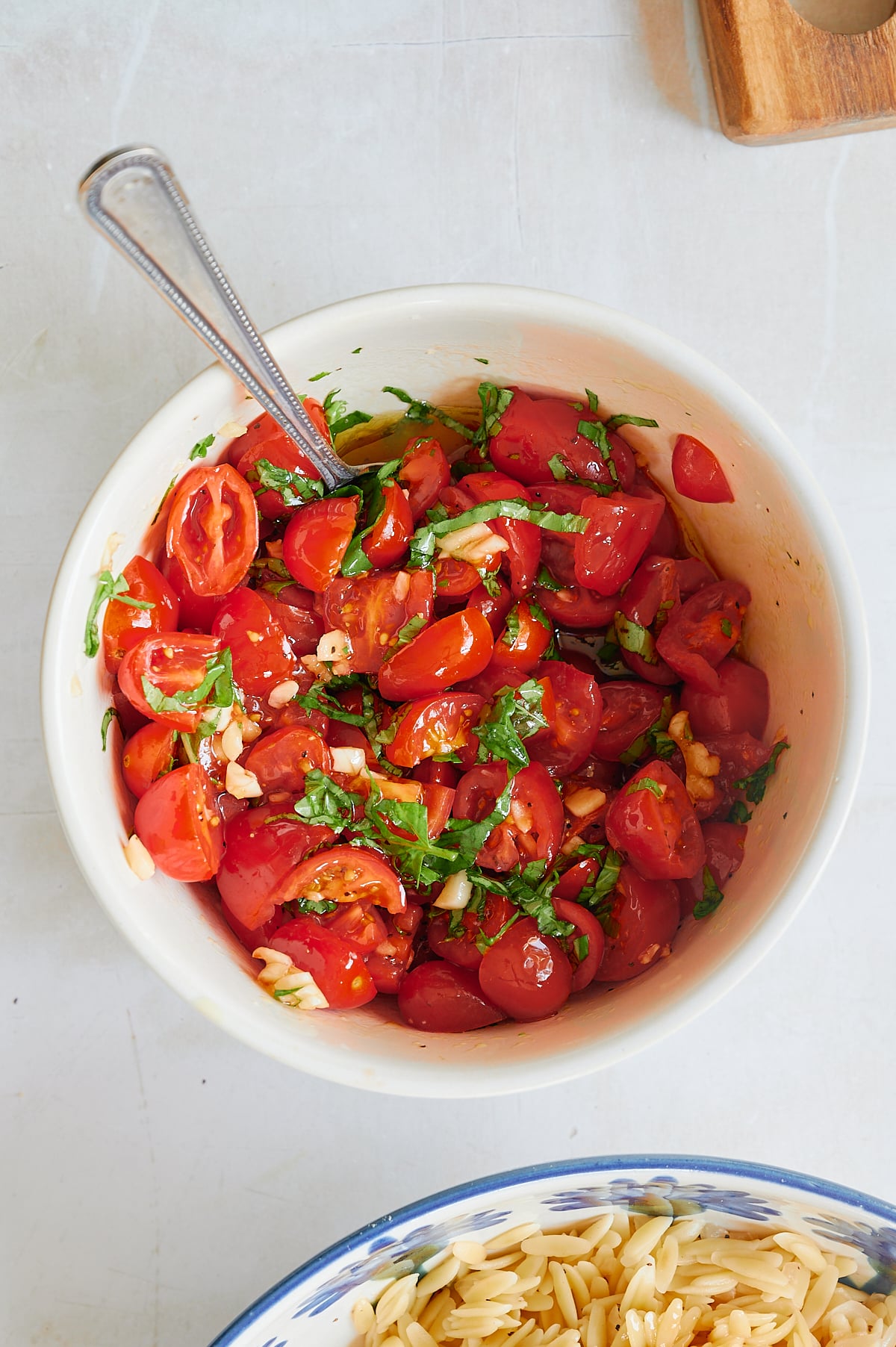 A white bowl of halved cherry tomatoes tossed with chopped fresh basil, garlic, and oil, with a spoon resting inside.